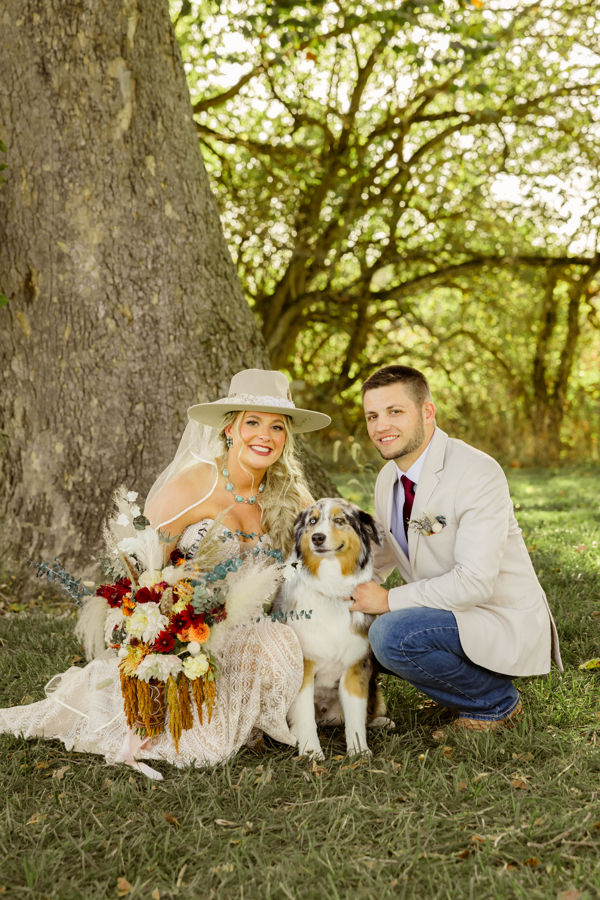 Rustic Columbus wedding venues A couple in formal attire kneels beside a dog under a large tree. The woman holds a colorful bouquet, and both smile in a sunny, lush setting.