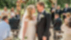 Barn Wedding Venue Ohio. Bride and groom smiling at each other under a floral arch in an outdoor wedding setting. Guests seated, greenery in the background, sunny day.