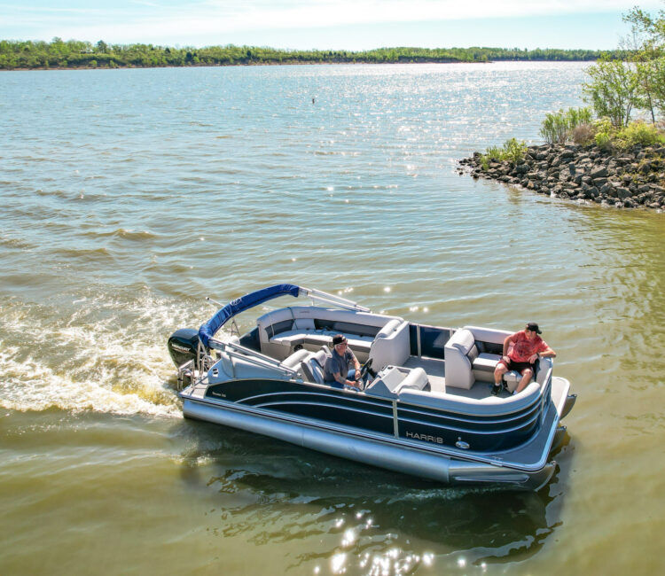 Ohio Wedding Venue. A blue pontoon boat with two people glides on a lake under a clear sky, near a rocky shore. The boat has "Harris" on its side.