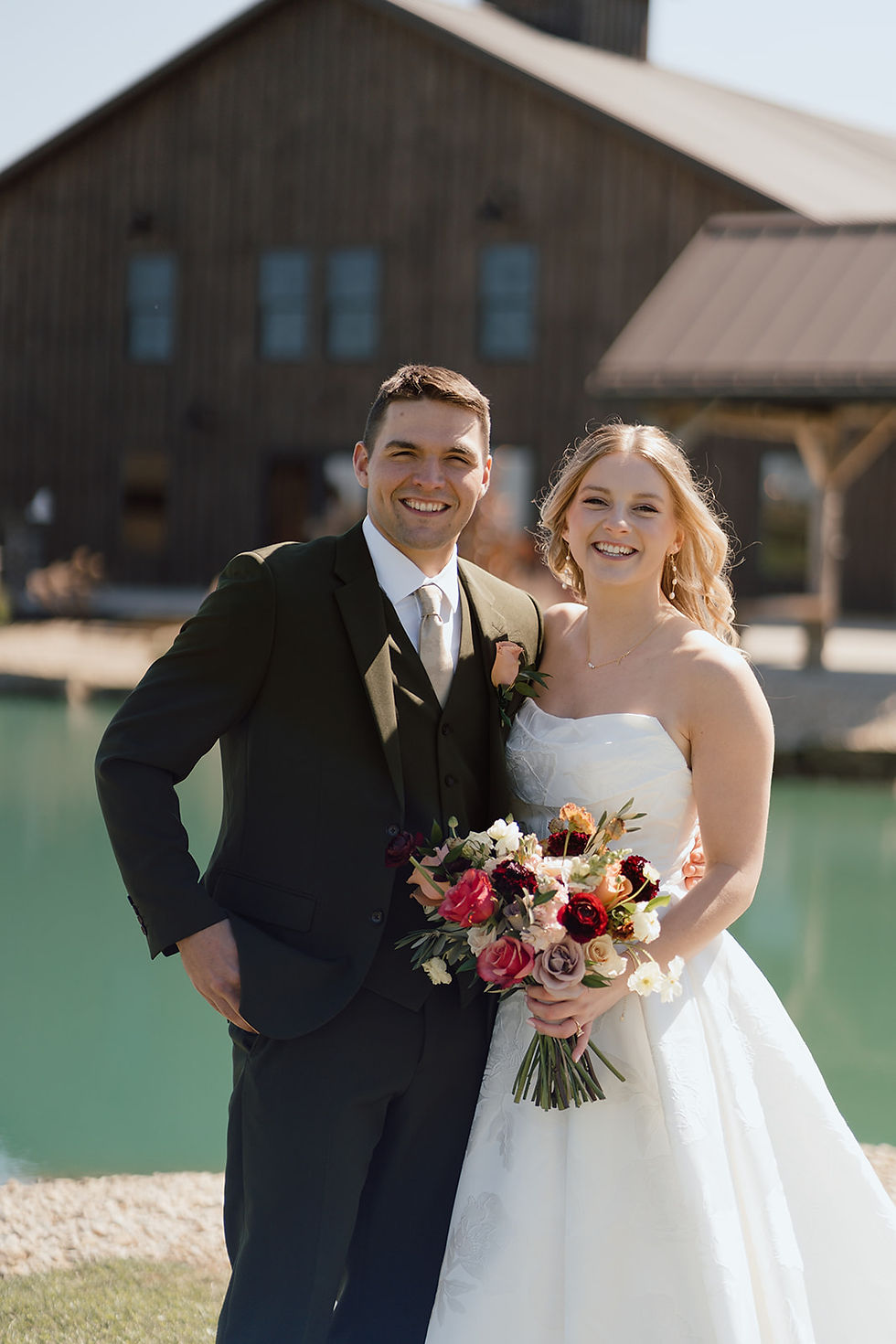 Central Ohio Wedding Venue Smiling bride and groom in wedding attire pose by turquoise water in front of a barn, holding a colorful bouquet.