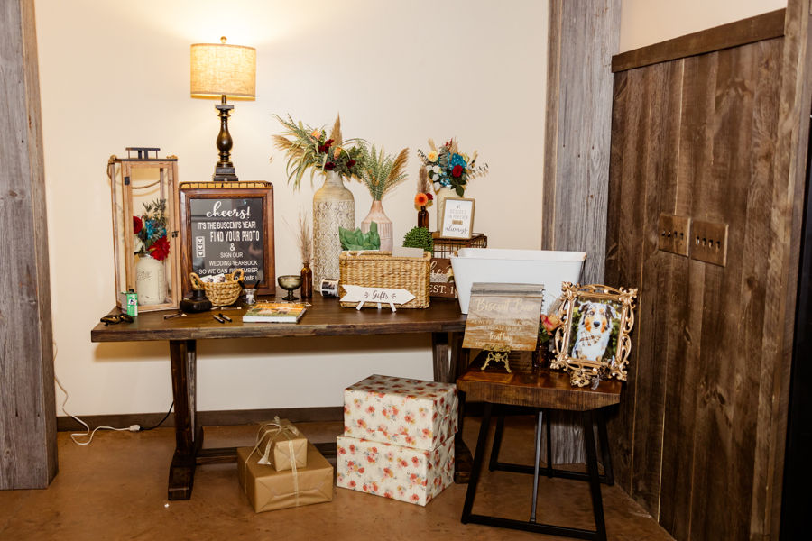 Barn Wedding Venues in Ohio. Gift table with vintage decor, framed dog photo, sign reading "cheers," flowers, and wrapped boxes. Warm lighting and rustic wood tones.