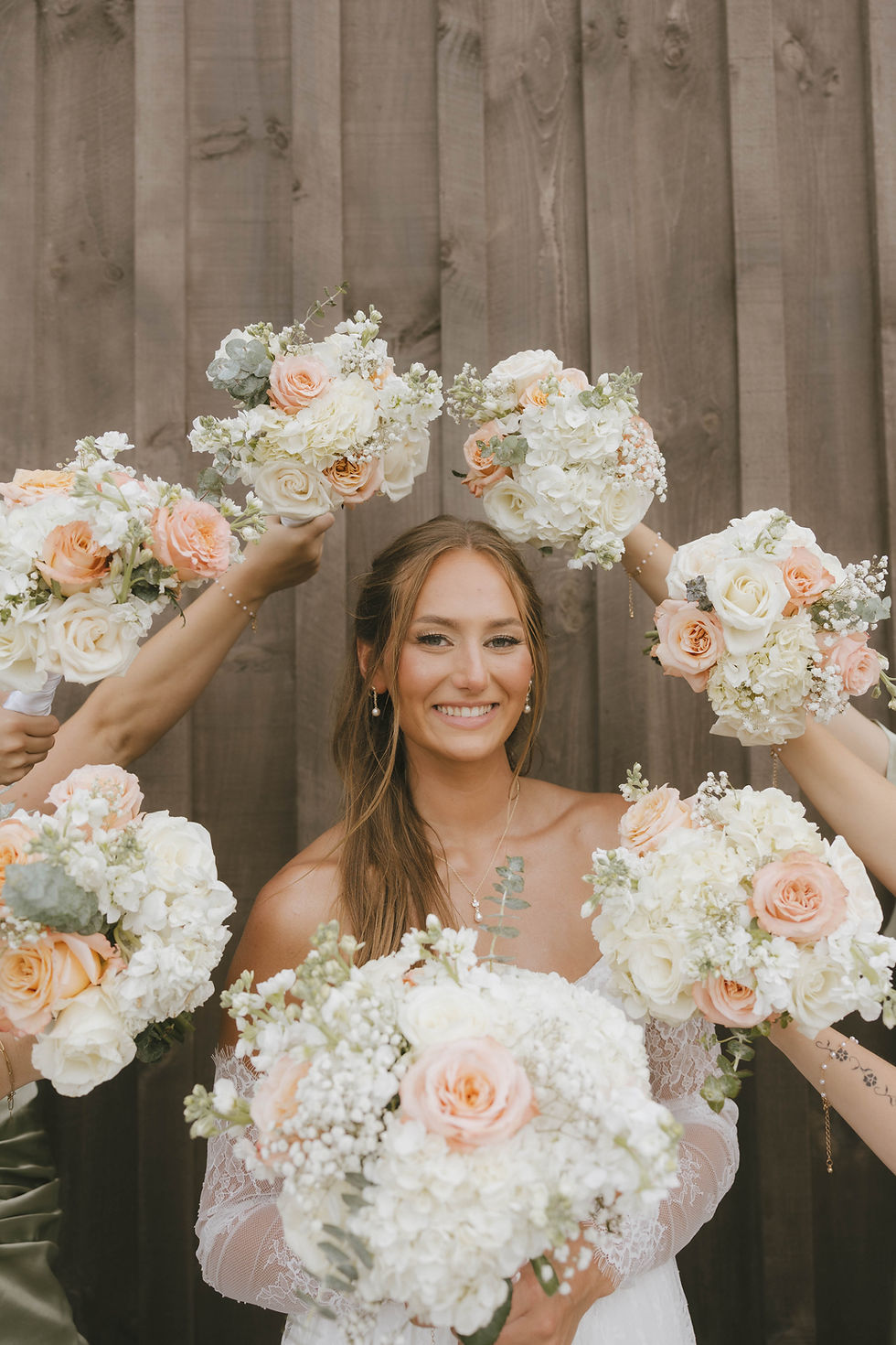 Grove City, OH Wedding Venue Smiling bride in white lace dress stands before wood fence as hands hold peach-and-white flower bouquets around her head