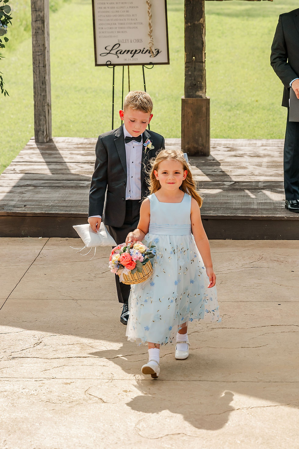 Barn Wedding Venues in Ohio. Young boy in a suit and girl in a blue dress walk on a sunny patio. Girl holds a basket of flowers; sign reads "Bailey & Chris Lamping."