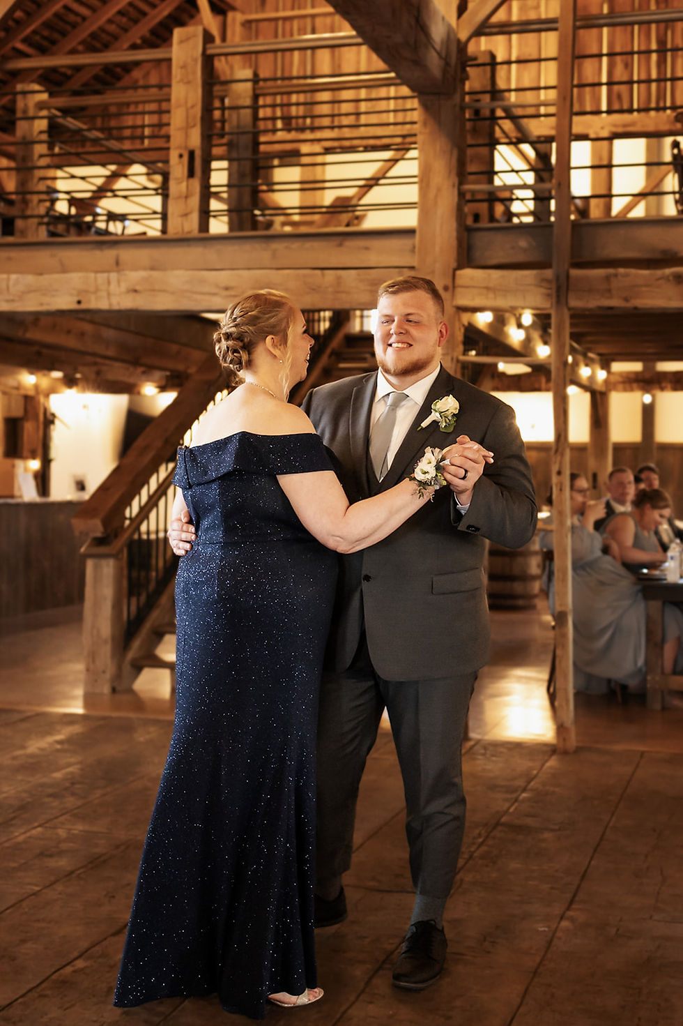 Barn Wedding Venues in Ohio. A man in a suit and woman in a navy dress dance in a rustic barn setting, lit warmly. Guests seated in the background. Joyful mood.