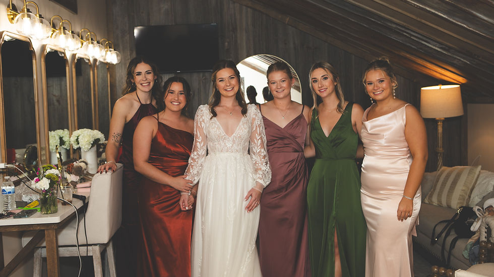 Barn Wedding Venues in Ohio. Six smiling women in formal dresses pose together in a wood-paneled room with vanity mirrors and lights, exuding a joyful atmosphere.