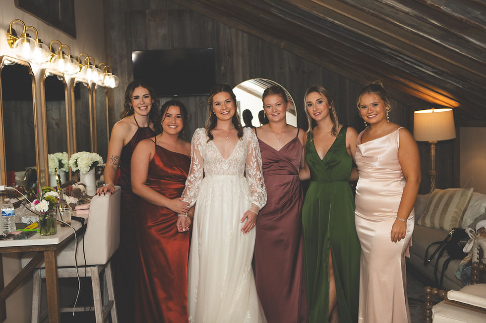 Barn Wedding Venues in Ohio. Six smiling women in formal dresses pose together in a wood-paneled room with vanity mirrors and lights, exuding a joyful atmosphere.
