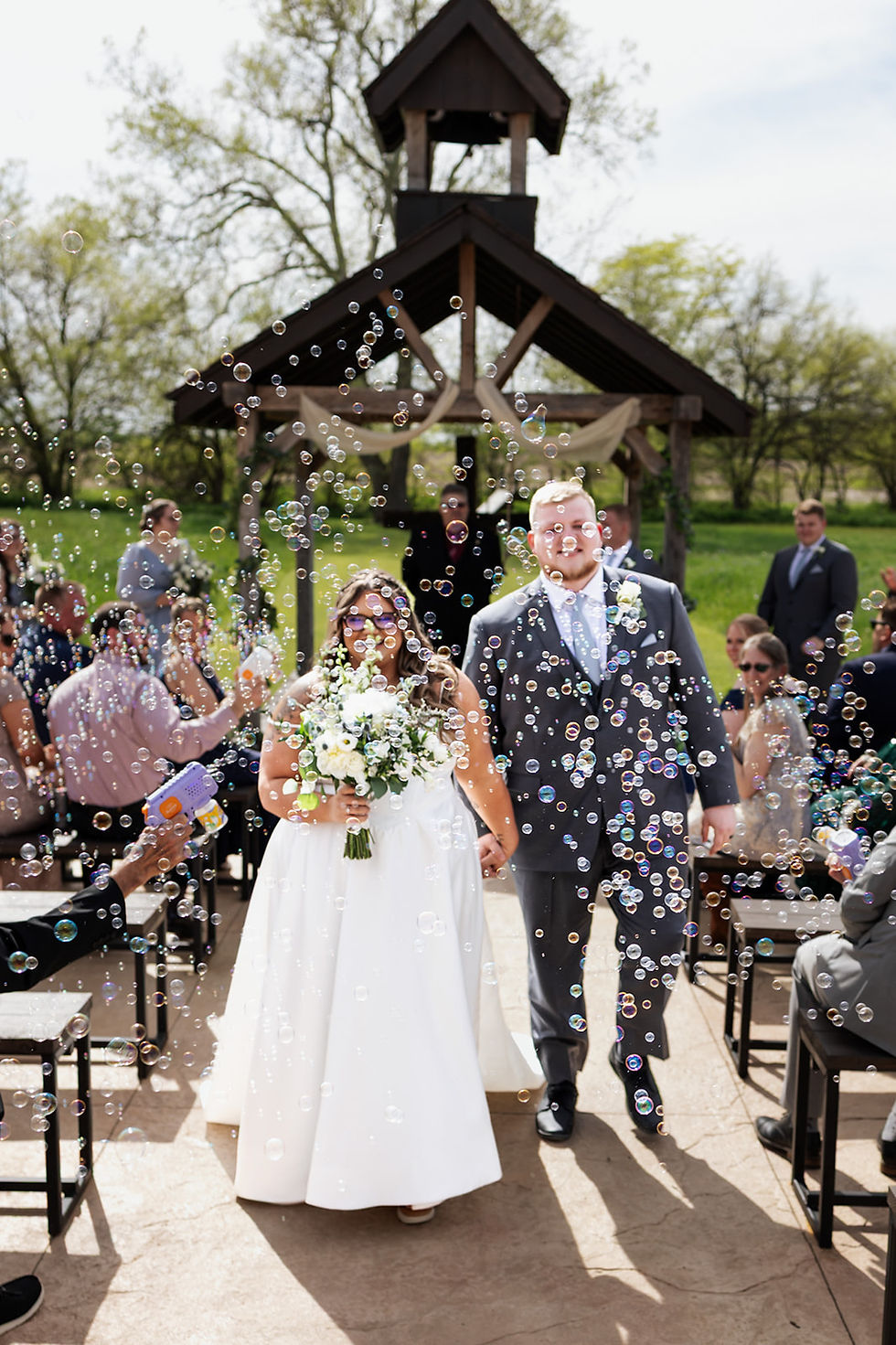 Ideas for Memorable Wedding Experience Ohio. Bride and groom smiling, holding hands, walk down an outdoor aisle with bubbles and guests in a scenic setting. Bride holds white bouquet.