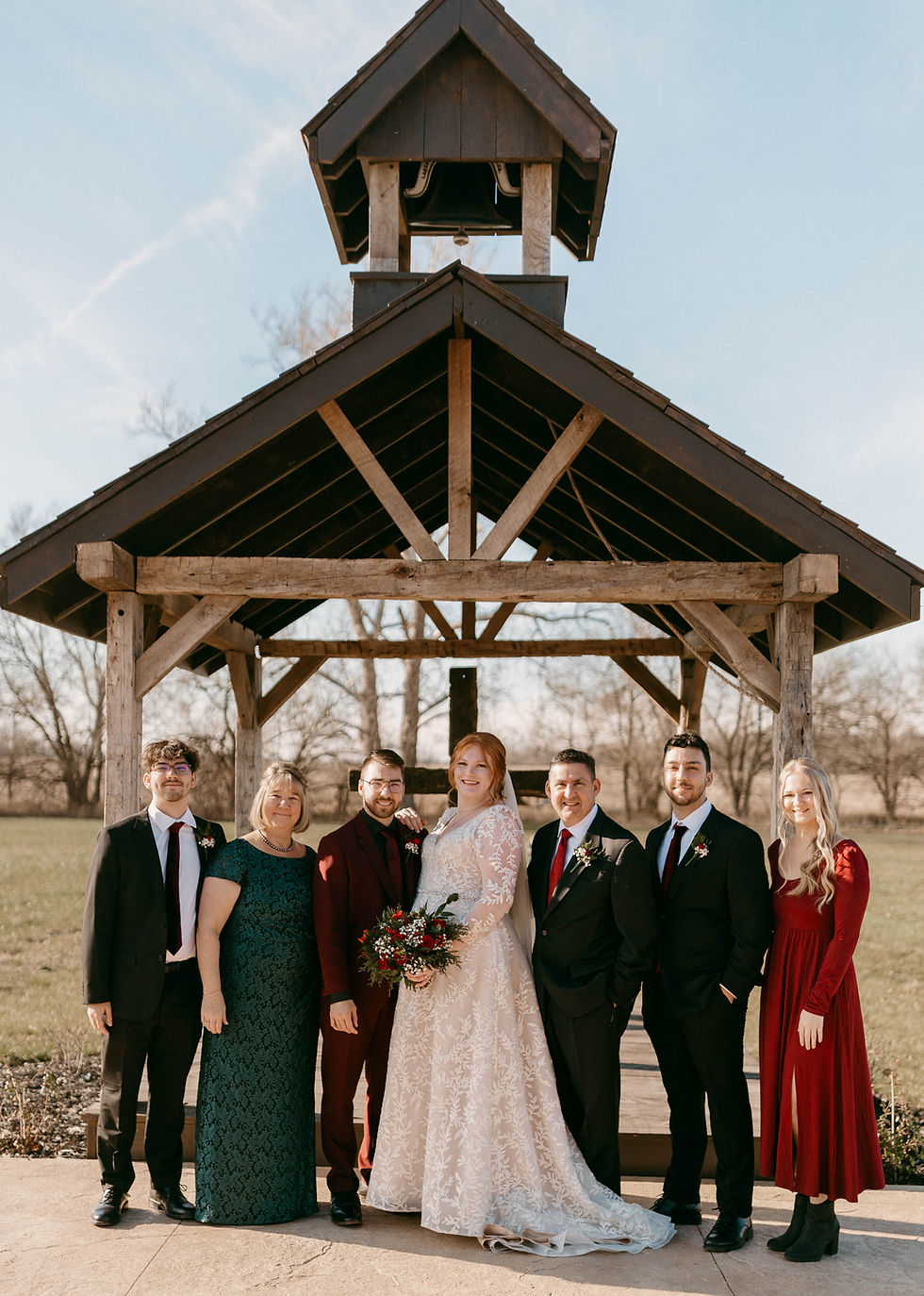 Barn Wedding Venues in Ohio. A newlywed couple poses with family under a rustic wooden structure. The bride holds a bouquet. Everyone is smiling in formal attire outdoors.
