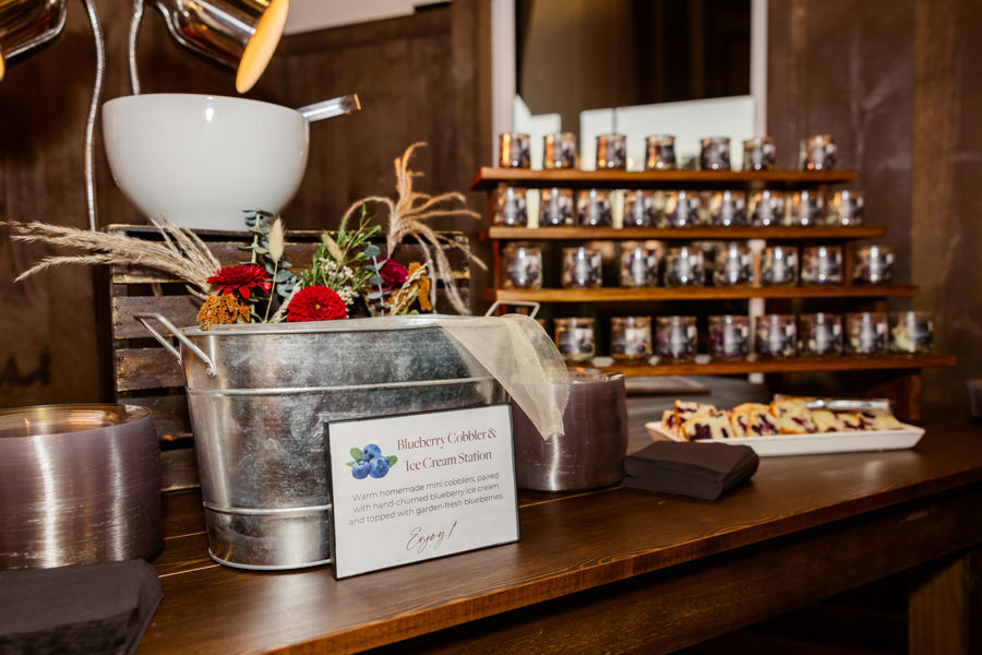 wedding venue Ohio Blueberry Cobbler & Ice Cream Station with a metal tub holding flowers. A sign describes the setup. Rows of jars and dessert display nearby.