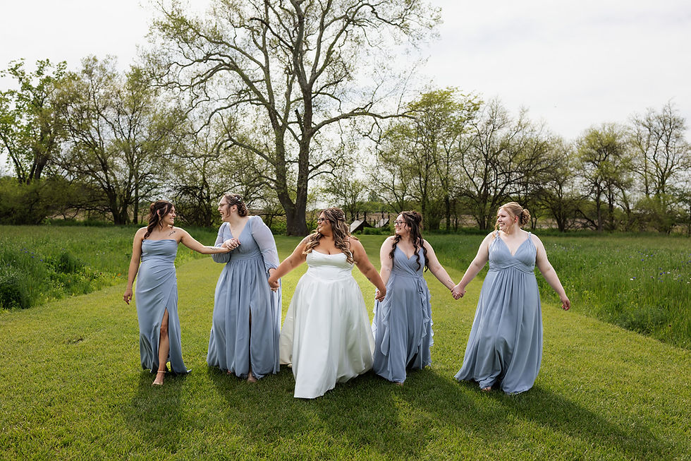Barn Wedding Venues in Ohio. Five women in gowns walk hand-in-hand on grassy field, with trees in the background. The scene is joyful and vibrant.
