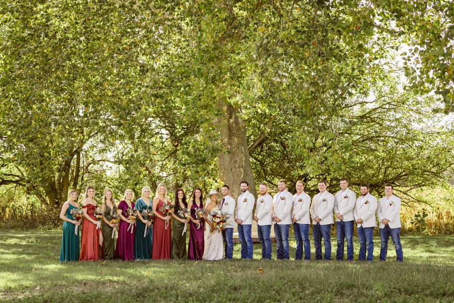 Ohio Wedding Venues A wedding party stands outdoors under a large tree. Bridesmaids wear colorful dresses, holding bouquets; groomsmen in white shirts and jeans.