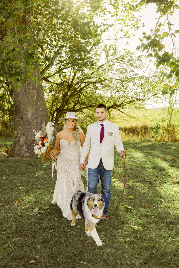 Rustic Columbus wedding venues A couple in wedding attire, holding hands in a sunny garden, walks with a cheerful dog. The woman holds a colorful bouquet.