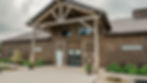Barn Wedding Venue Ohio. Wooden barn with a metal roof, stone accents, and greenery. A sign with a bow is at the entrance. Cloudy sky in the background.