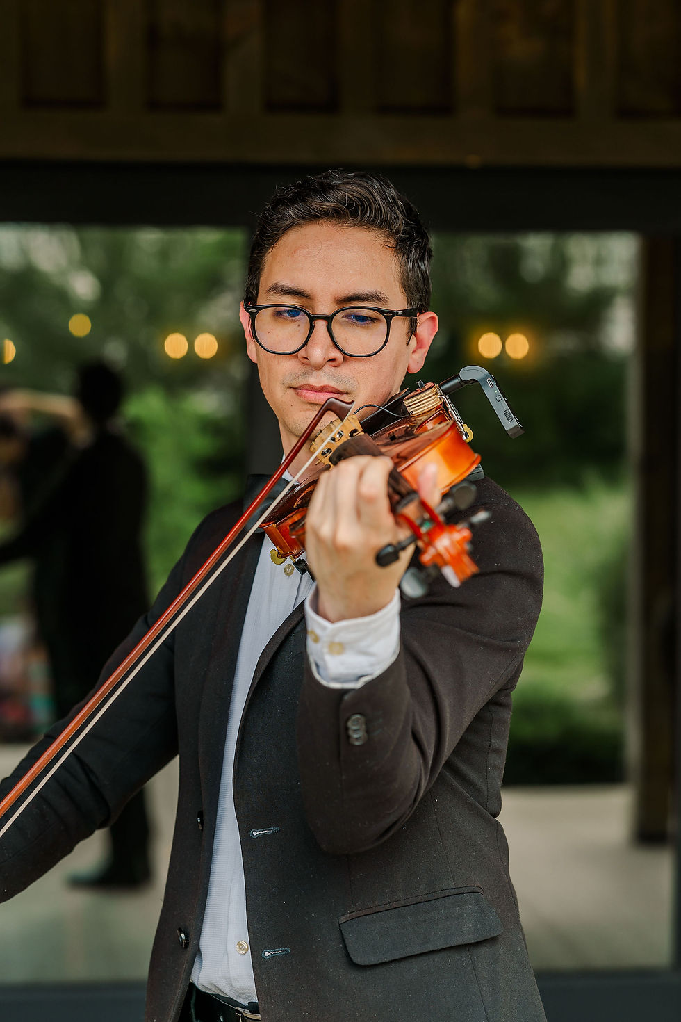 Pickaway County Wedding Venue Young man in a black suit plays violin outdoors, focused, with blurred greenery and warm lights behind him.