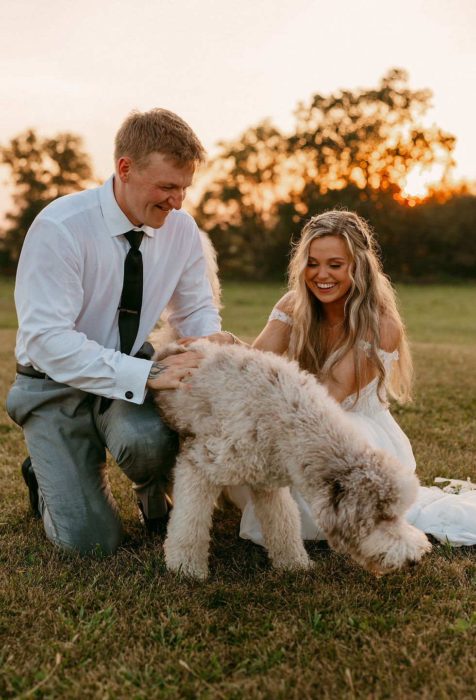 Rustic Columbus wedding venues A couple in formal attire smiles at a fluffy dog on a grassy field during sunset. Trees are visible in the background, creating a warm glow.