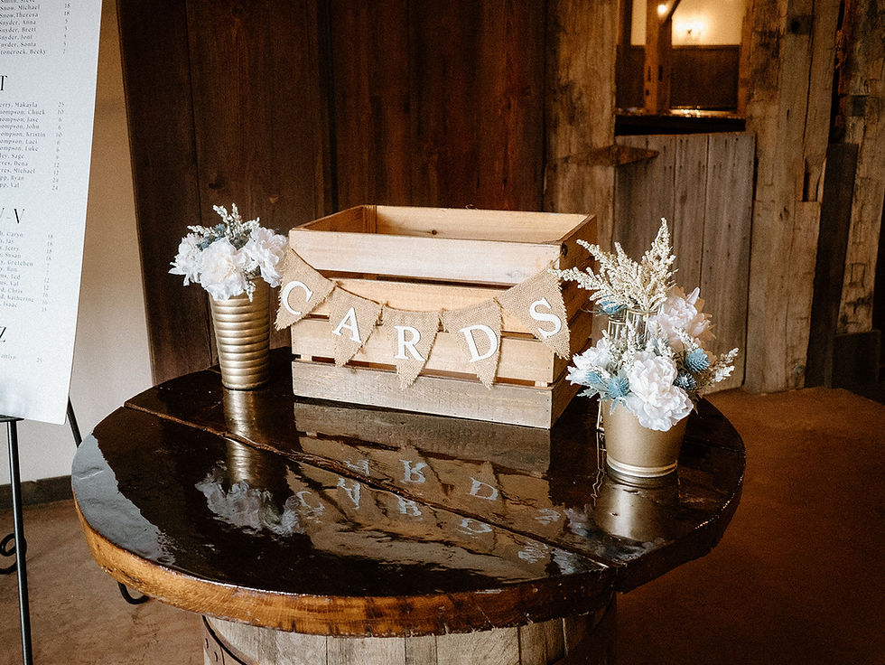 Barn Wedding Venues in Ohio. Wooden card box with "CARDS" bunting on rustic table, flanked by white flower arrangements in gold vases. Warm, wooden background.