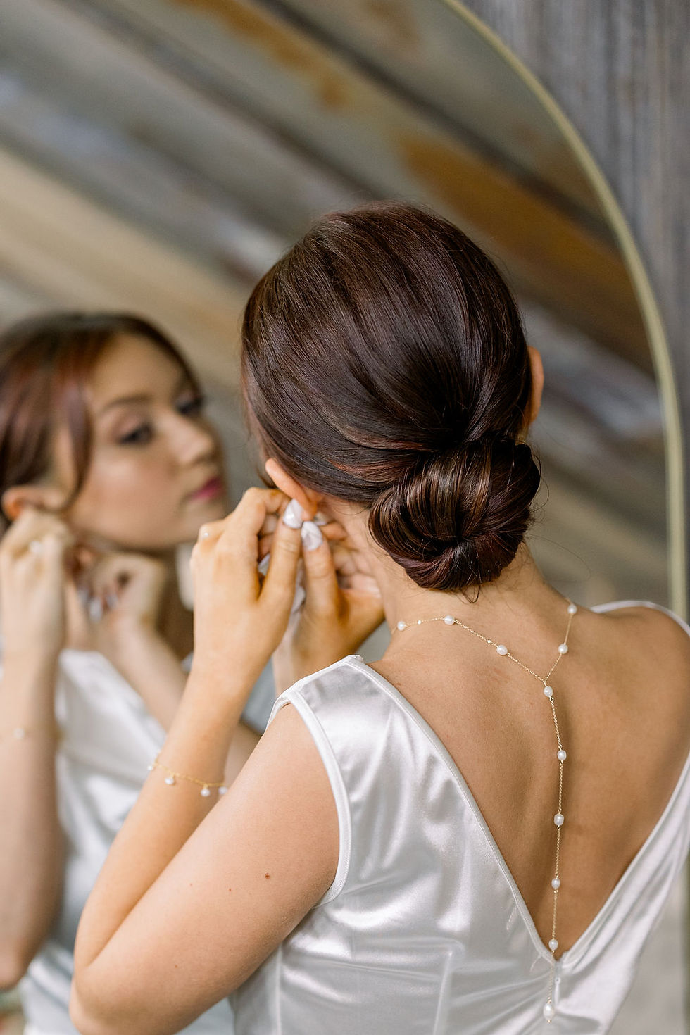 Central Ohio Wedding Venue Woman in a white satin dress adjusts her earring in a mirror, wearing a pearl-back necklace in a soft bridal prep scene.