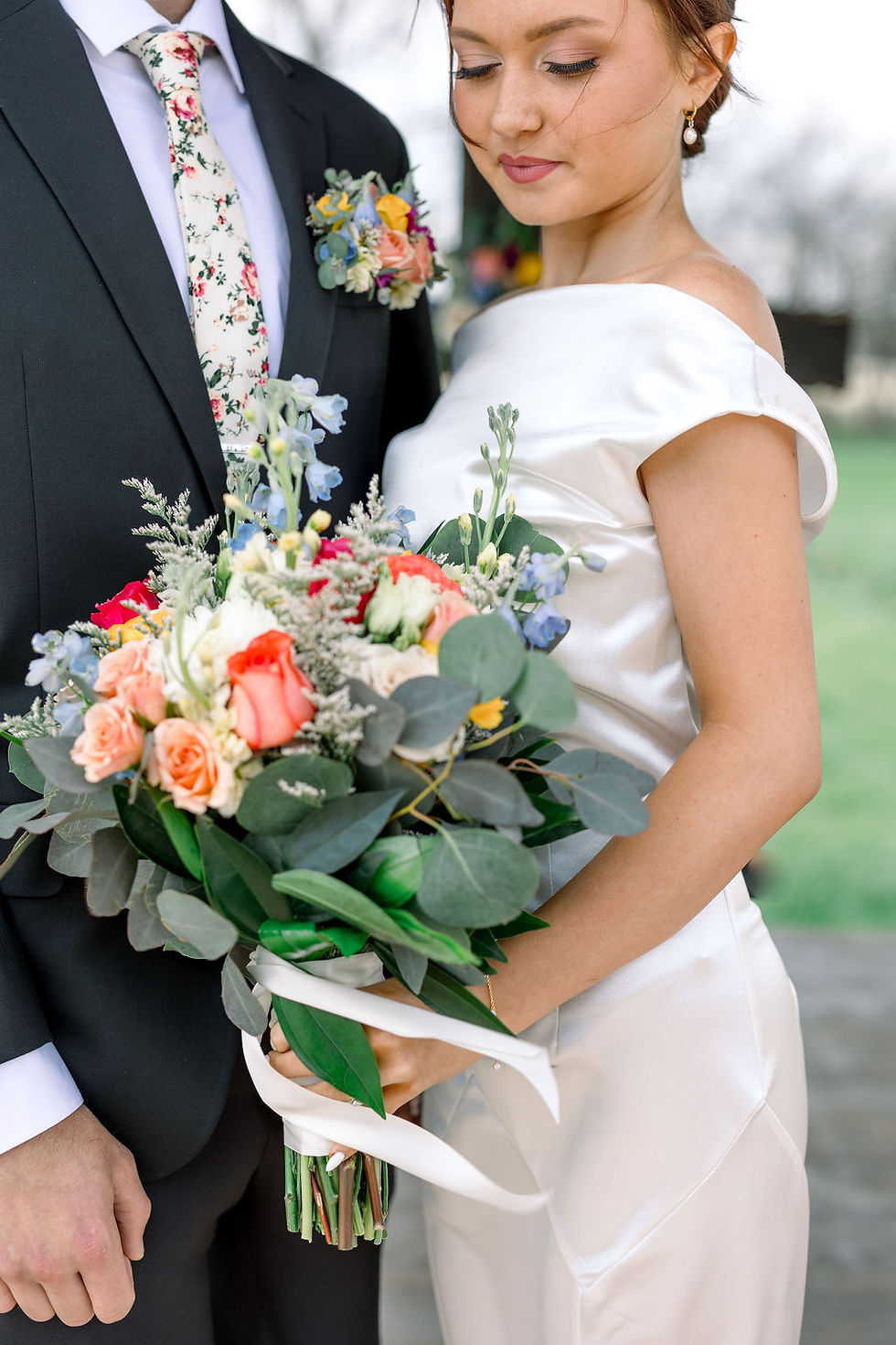 Barn Wedding Venues in Ohio. Bride with bouquet of colorful flowers stands beside groom in black suit with floral tie. Soft focus outdoor background, serene mood.