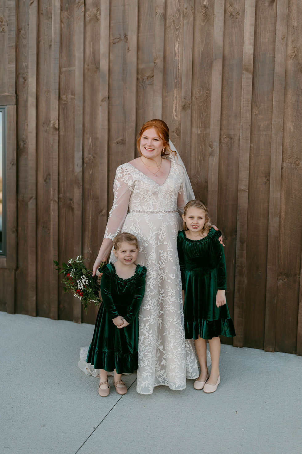 Barn Wedding Venues in Ohio. Bride in white lace dress holds bouquet, smiles with two girls in green dresses against wooden wall, creating a joyful wedding scene.