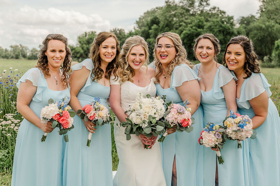 Barn Wedding Venues in Ohio. Bridesmaids in light blue dresses and the bride smile, holding colorful bouquets, in a lush outdoor setting with greenery.