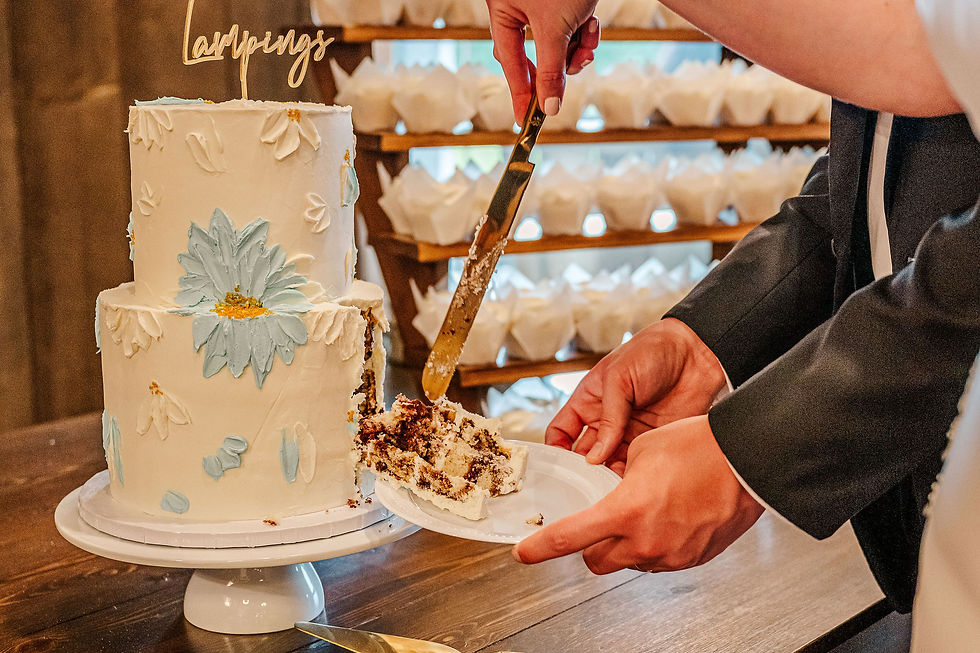 Barn Wedding Venue Ohio. Hands cut white cake with blue flowers; "Lamping" topper. Cupcakes in background suggest a celebration setting.