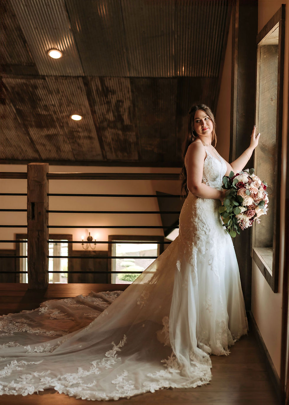 Barn Wedding Venues in Ohio. Bride in white gown holds bouquet, gazes out rustic window. Wooden interior, serene mood, soft lighting highlights lace train.