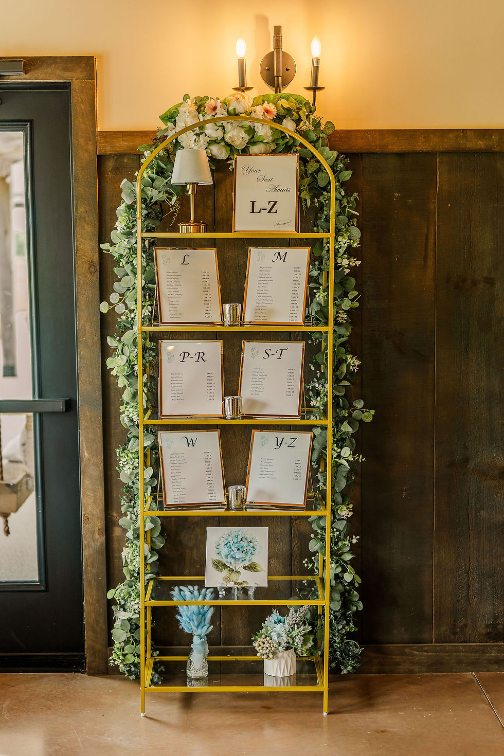 Barn Wedding Venues in Ohio. Wedding seating chart setup on a yellow shelf, decorated with greenery and flowers. Labels with letters L-Z in a warm, wooden room.