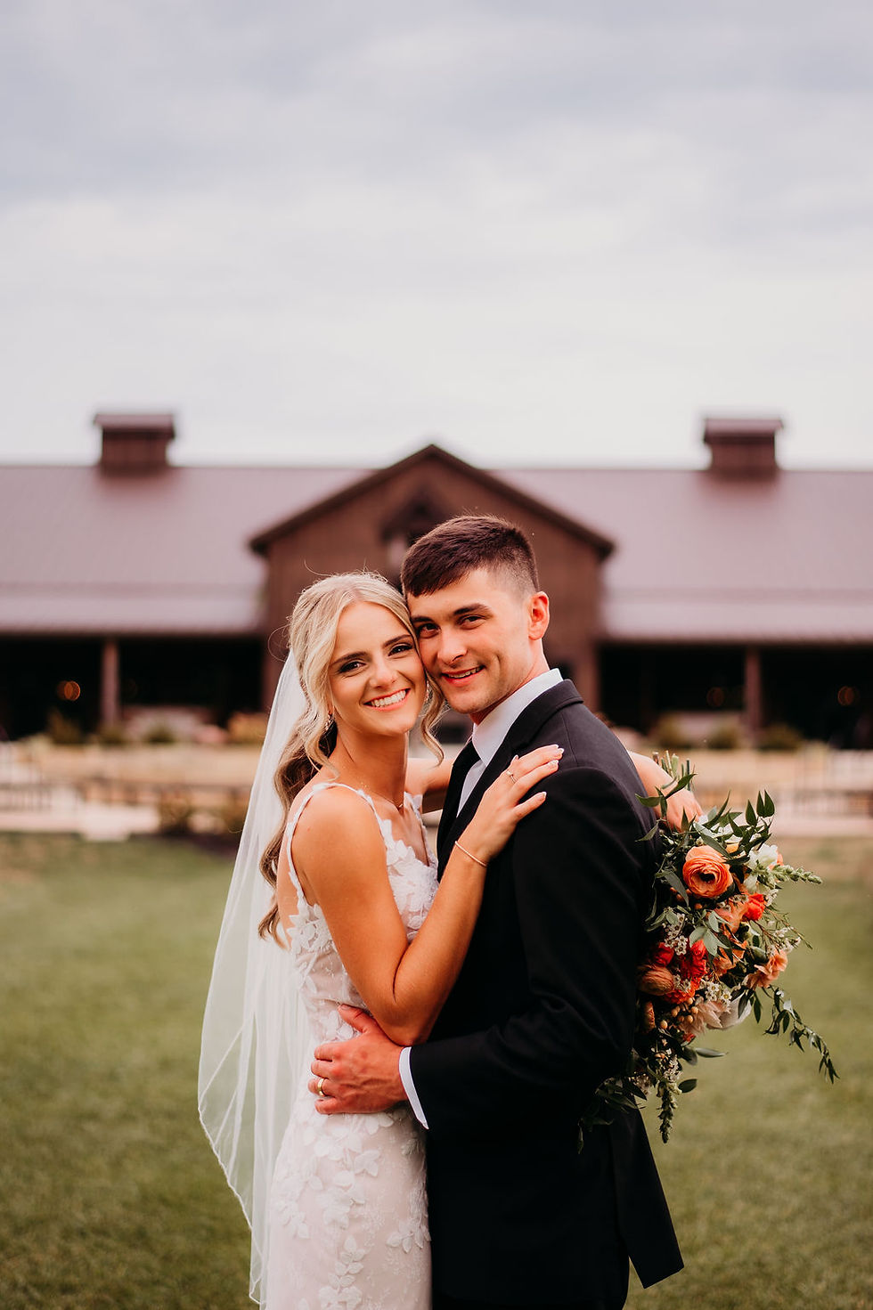 Wedding Guest List Bride and groom smiling, embracing outdoors. Bride in lace dress and veil, groom in black suit holding bouquet. Barn in background, overcast sky.