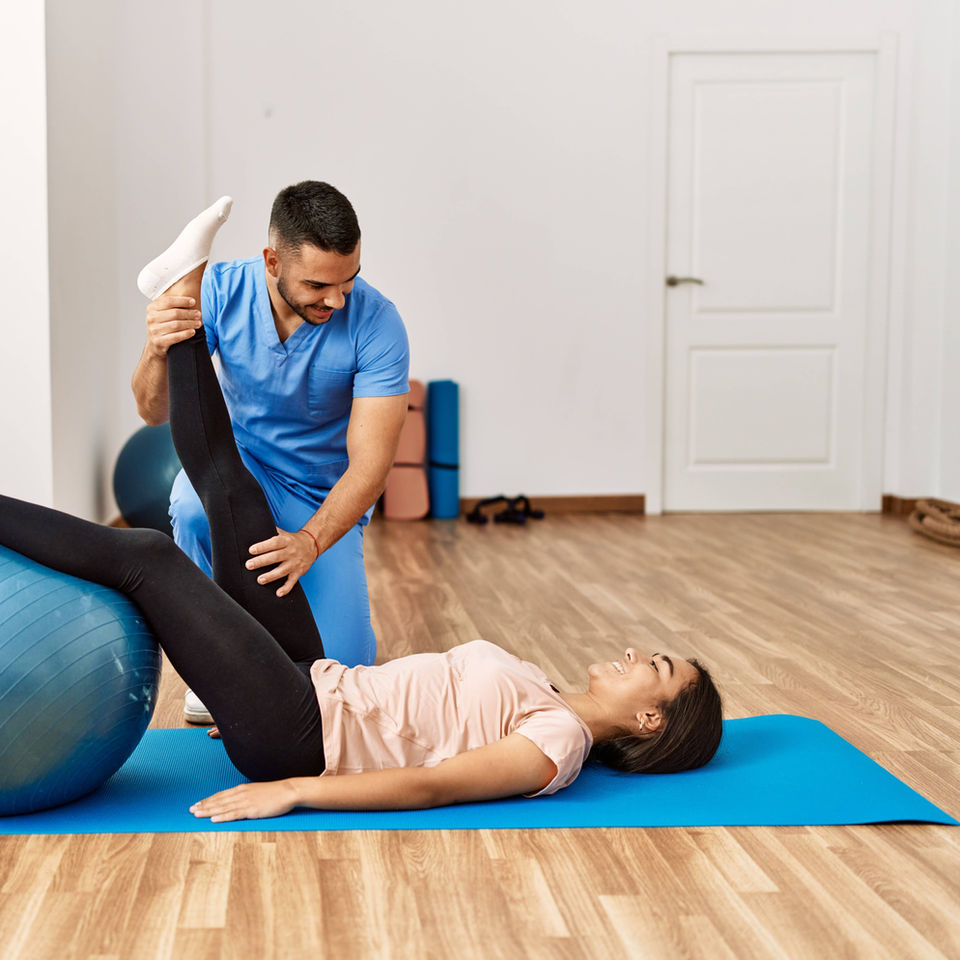 White male assisting a white female with stretches on a swiss ball.