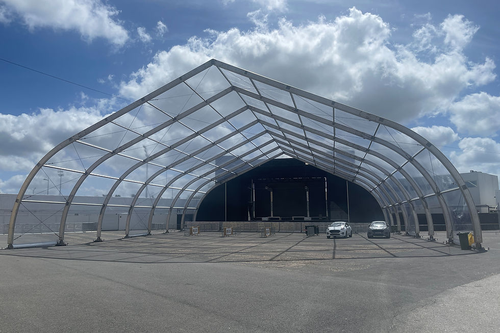 A large transparent event tent structure under construction, with two cars parked inside and a cloudy sky above.