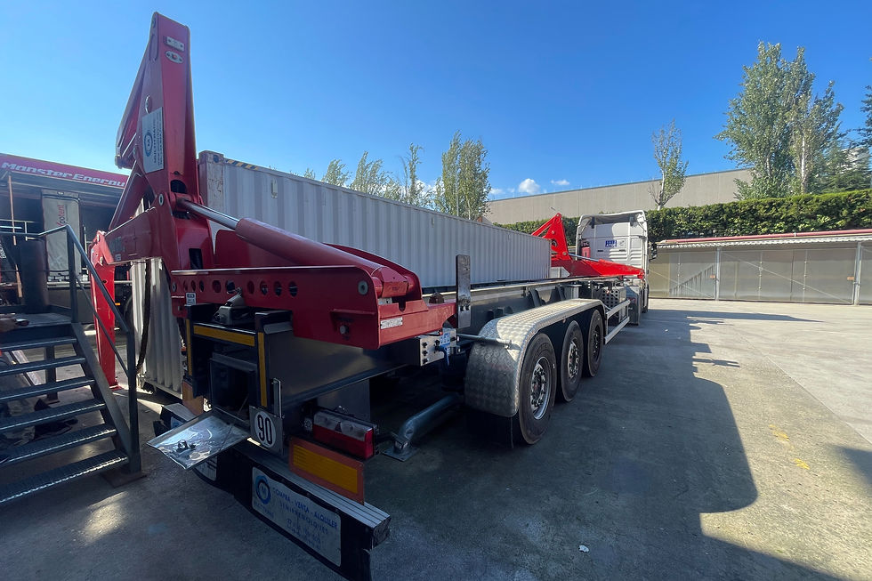 A side-loader trailer with its red lifting arms extended, positioned to move a white shipping container inside an industrial yard on a sunny day.