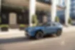 Blue SUV driving past modern buildings on a city street. The vehicle is in motion, with blurred background. Sunny day with clear skies.