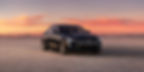 Black cadillac on a desert road during sunset, with a vibrant sky and distant mountains. The car's grille has a logo. Calm, open setting.