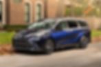 Blue Toyota minivan parked on a suburban street with a brick house in the background. Fallen leaves are on the ground beside it.