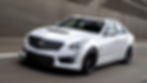 White Cadillac sedan driving on highway, blurred background. Sleek design with black wheels, grille, and visible badge. Dynamic motion.