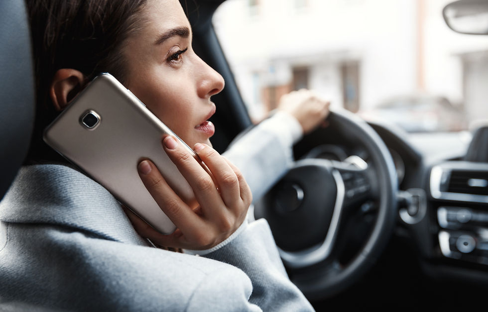 Woman in a gray coat drives a car while talking on a smartphone, looking distracted. Interior of the car visible.