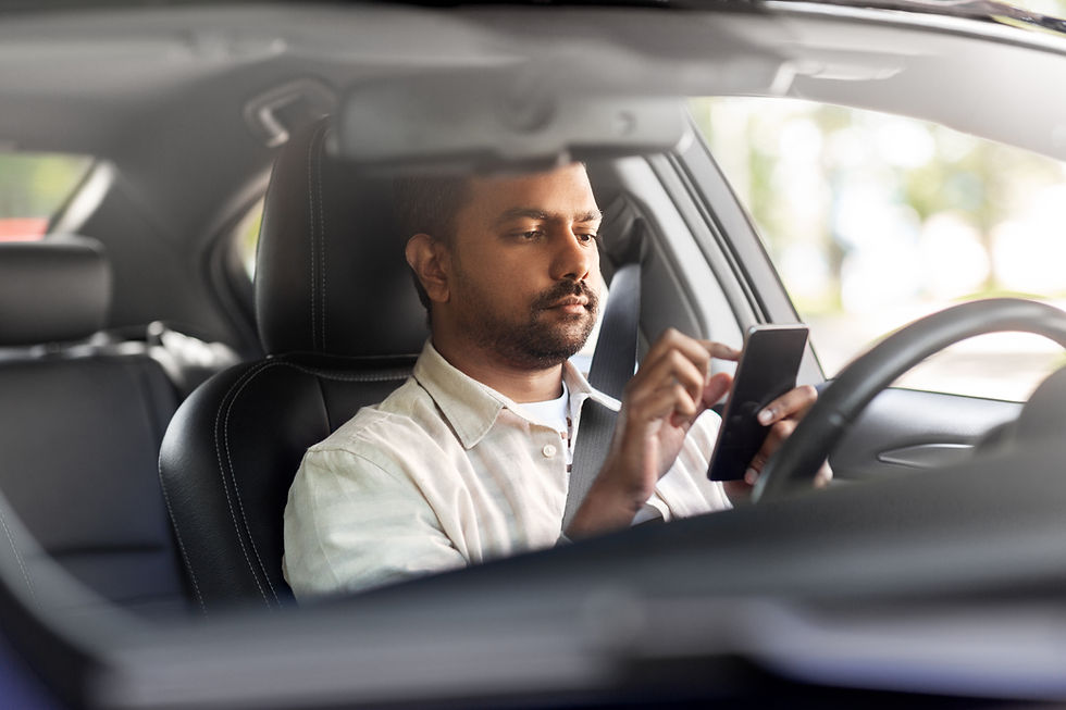 Man in white shirt uses smartphone to text while driving a car, with a focused expression. Blurred trees visible through window, interior is dark.