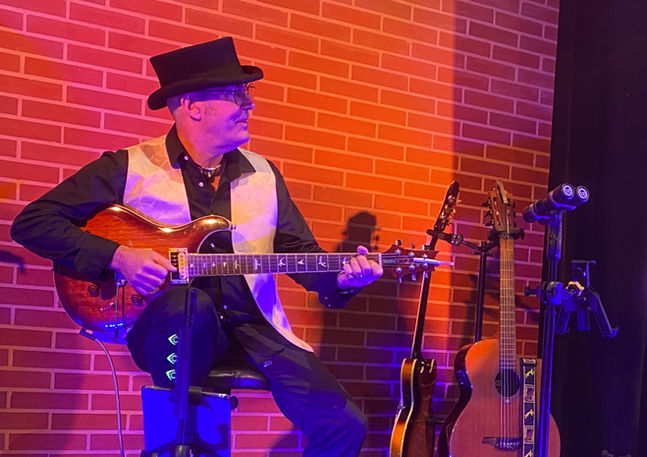Tommy Bird assis jouant de la guitare devant un décor de briques rouges pour l'agence Impulsion