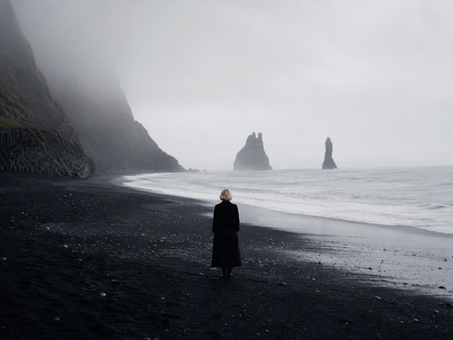 Lone woman in a long black coat stands on a foggy black-sand beach, facing sea stacks and cliffs.