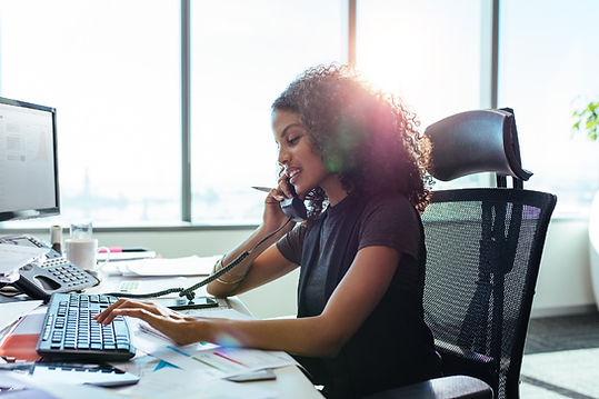 An attractive black woman with curley shoulder length hair, is seated at her computer in front of a blurry office window.