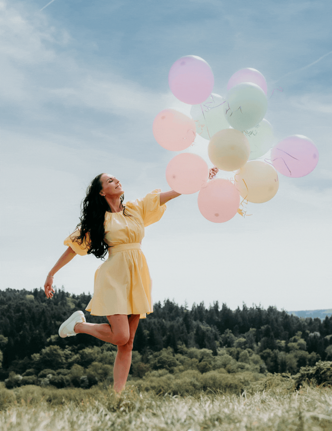 Gif images of a brunette female in a field wearing a buttercup yellow dress and holding a bunch of pastel coloured balloons.