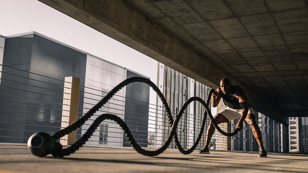 Man exercising with battle ropes under concrete structure, urban background, wearing black tank and white shorts, intense focus, sunlight.