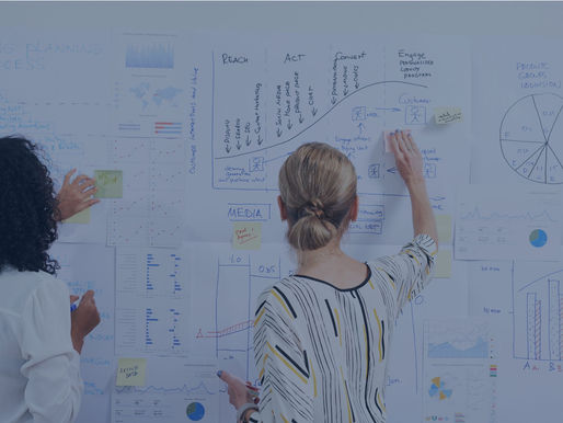 Two women collaborate on a project, sticking notes on a wall covered with charts, graphs, and the text "Marketing Planning Process."