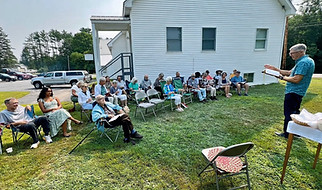 A group of people listening to a minister speaking outdoors, white building backdrop.