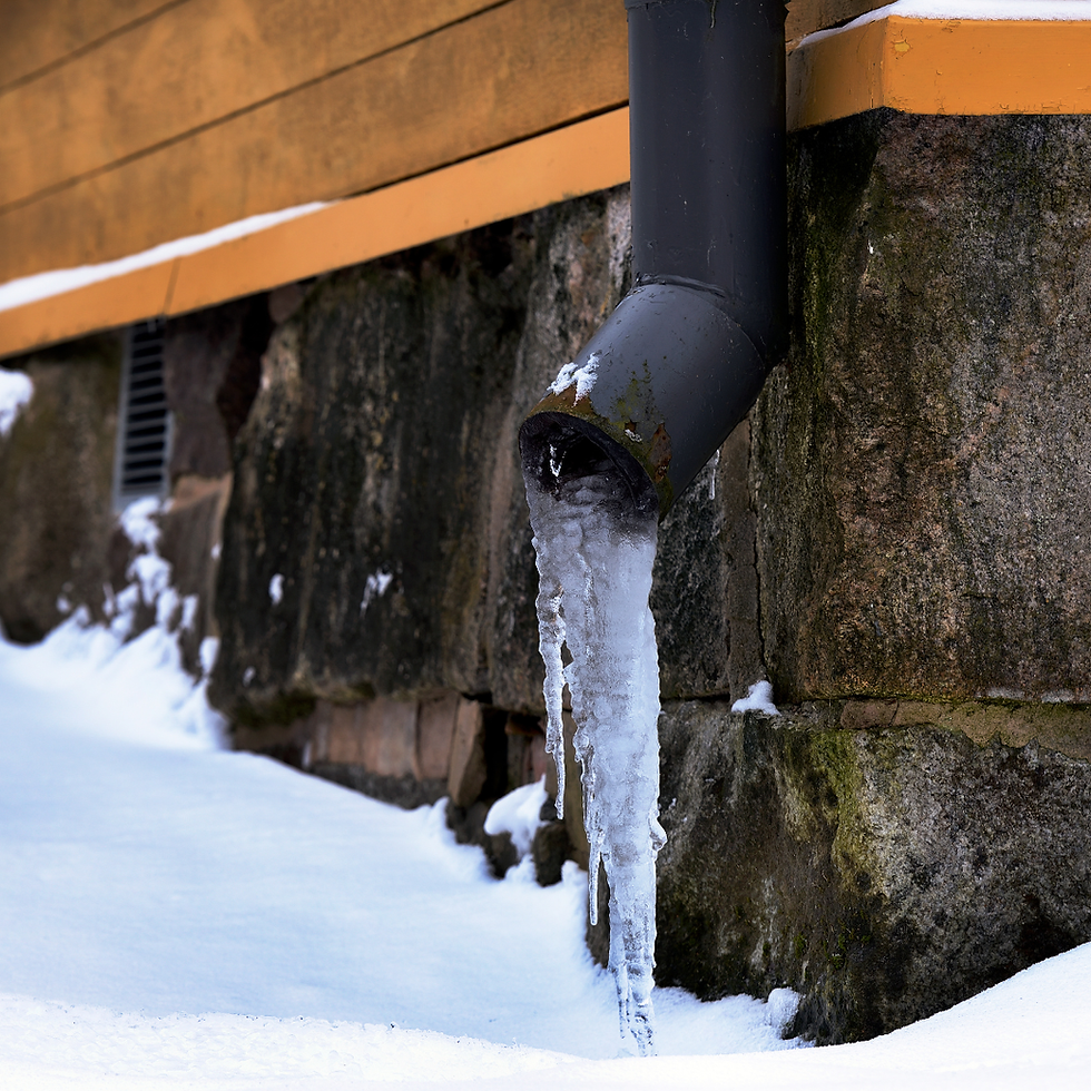 Icicles hang from a black downspout against a stone wall. Snow covers the ground, and the building's yellow trim adds contrast.