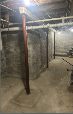 Dimly lit basement with concrete walls and floor, exposed pipes, and a wooden support beam. Sparse setting with a utilitarian feel.