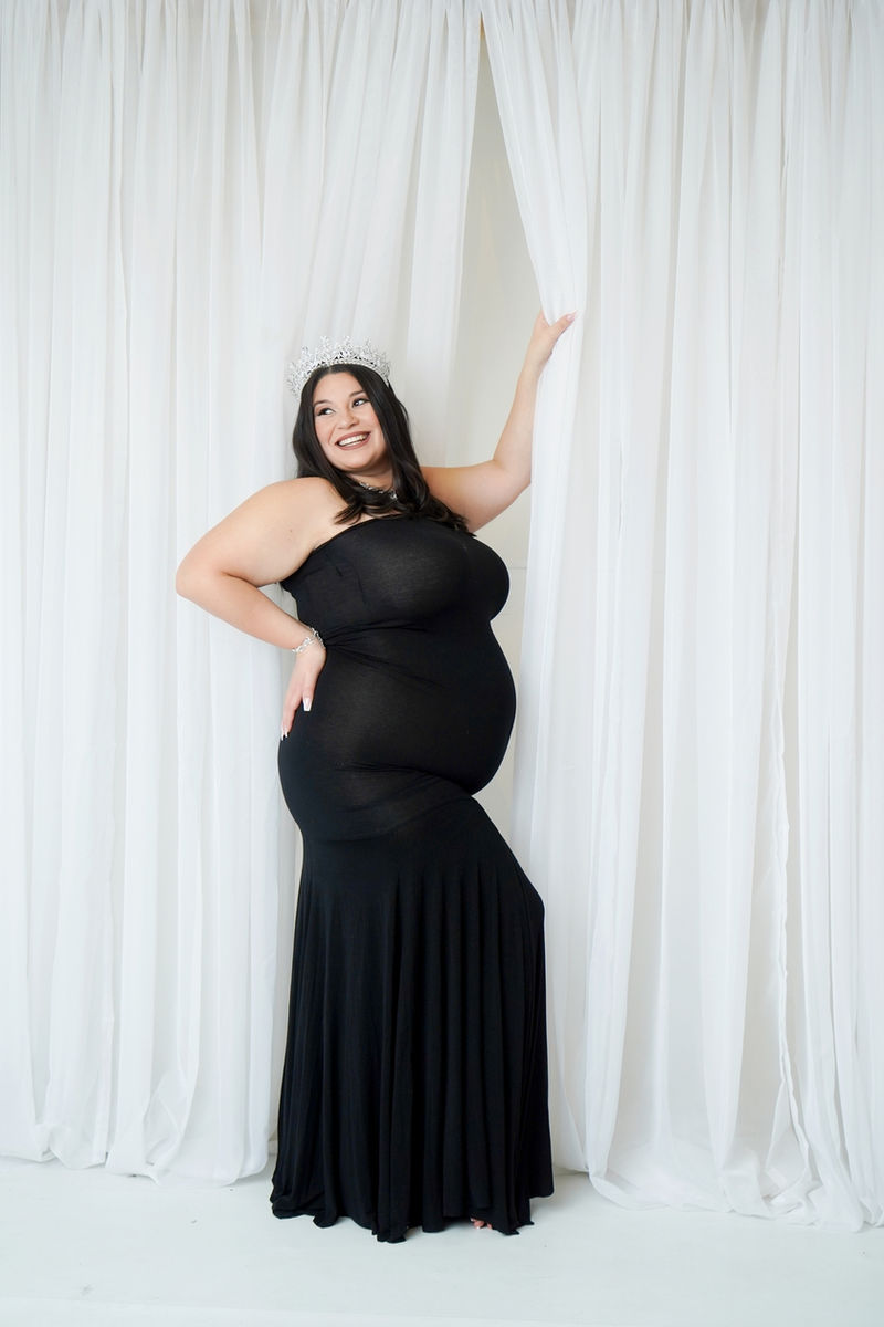 Expectant mother in a black strapless maternity gown and silver tiara smiling among sheer white drapes during a Folsom studio maternity session