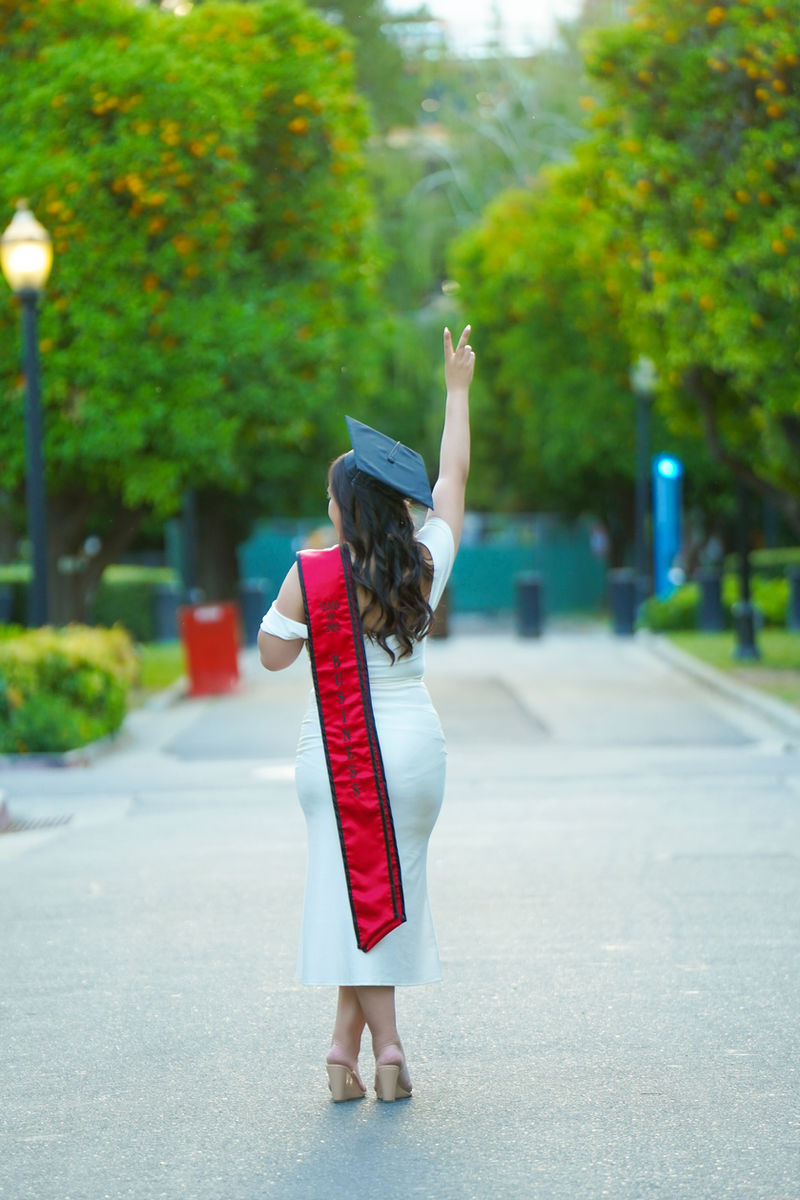 College graduate in a white dress and red sash walking down a tree-lined campus path during an on-location Sacramento graduation session