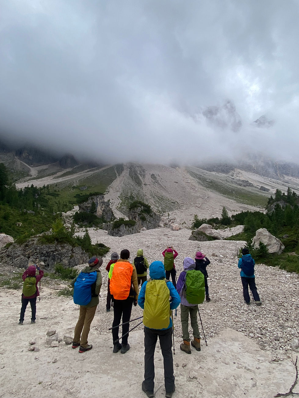 Hikers with colorful backpacks stand on a rocky trail, photographing a foggy mountain valley under low clouds.
