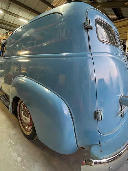 Technician performing paint restoration on a 1952 Chevy Panel Wagon, focusing on the classic design and weathered paint. The car is in a well-lit garage, with tools and restoration products nearby. The technician is carefully applying products to rejuvenate the vehicle's exterior, highlighting the dedication to restoring its vintage charm.